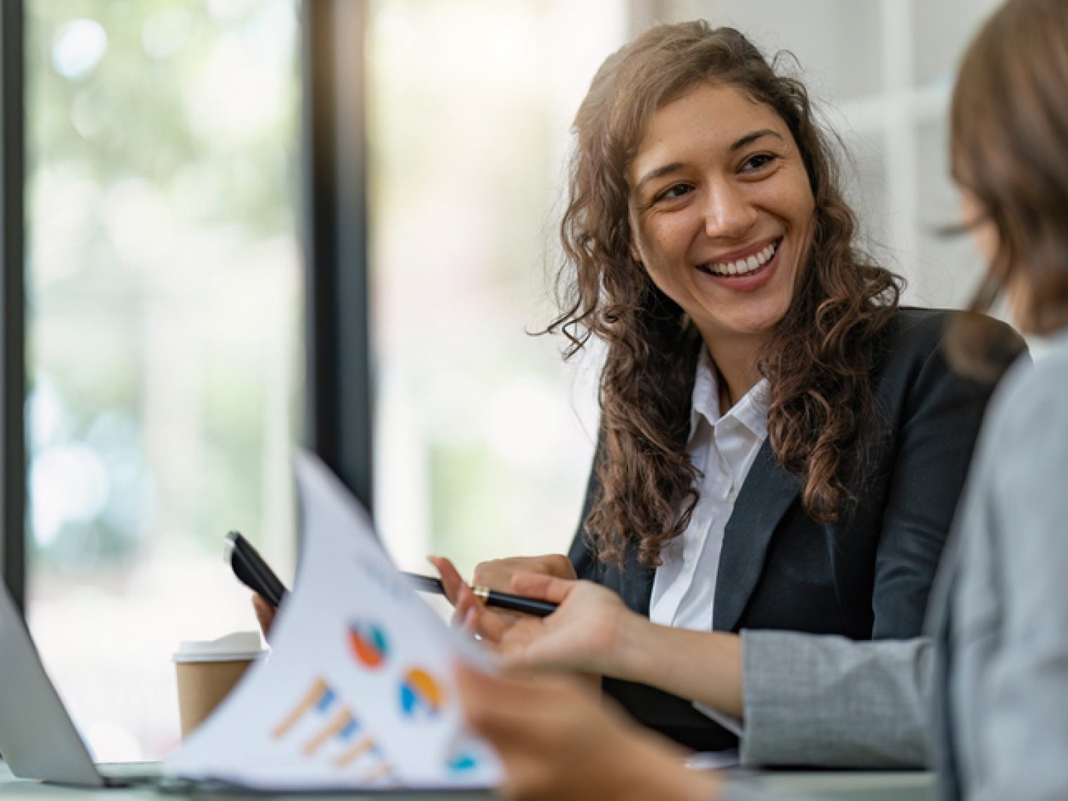 A smiling woman in business attire engaging in a friendly discussion with a colleague or client over documents and a laptop in a well-lit office setting.