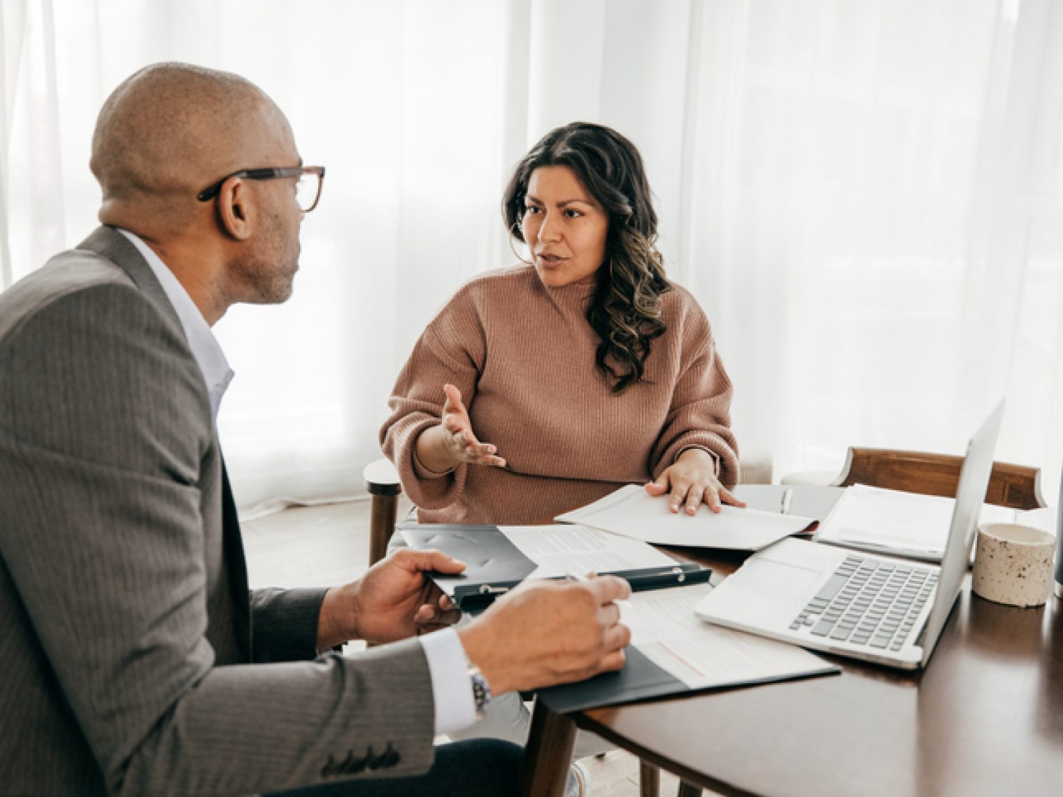 A focused conversation is taking place between a woman and a man sitting at a table with documents, a laptop, and a tablet, in what appears to be a serious professional consultation.