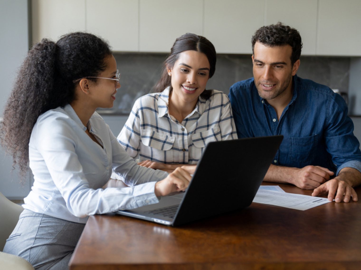 A professional woman is showing something on a laptop to a couple, who are looking on with interest, suggesting a consultation or meeting at a home or office.