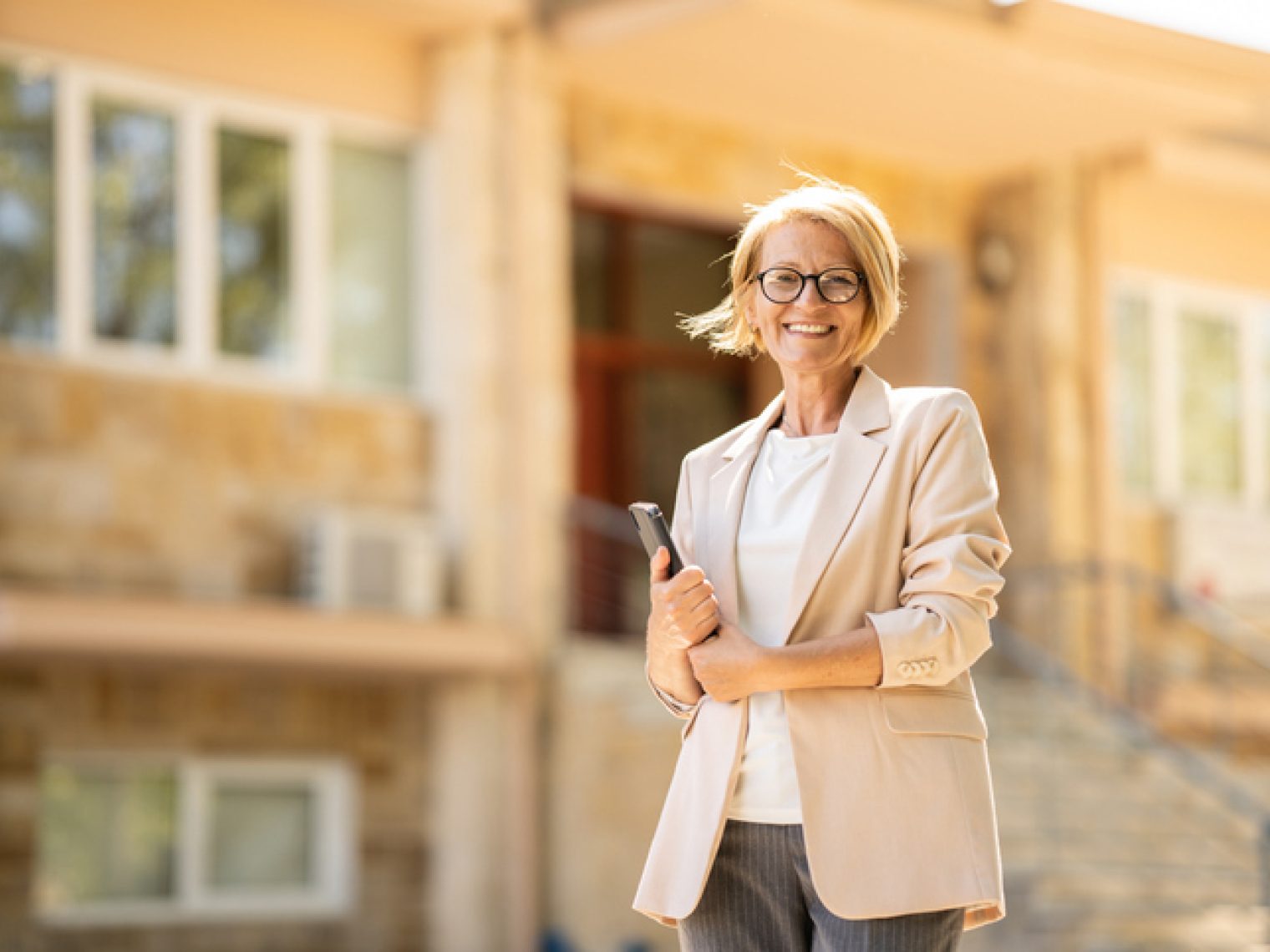 A confident woman in professional attire, holding a tablet, stands smiling in front of a building.