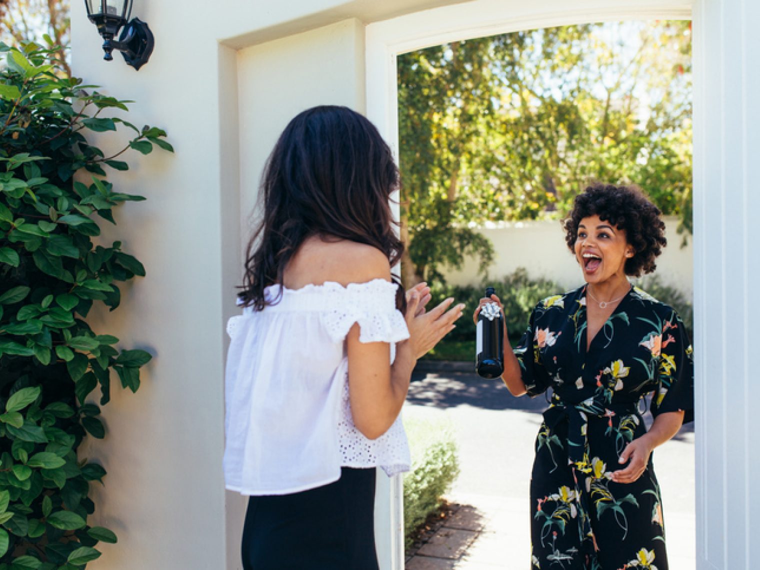 A woman in a floral dress excitedly presents a bottle to another woman at the doorway as a housewarming gift.