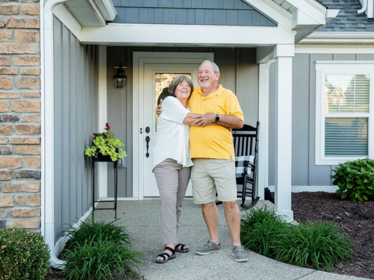 A happy elderly couple embraces in front of their home's entrance, smiling broadly in a welcoming suburban setting.