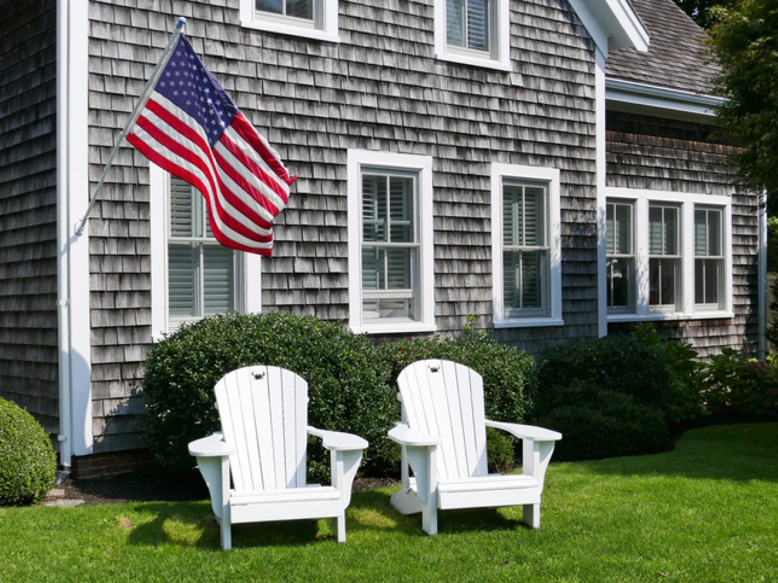 Two white Adirondack chairs on a well-kept lawn in front of a shingled home with an American flag displayed.