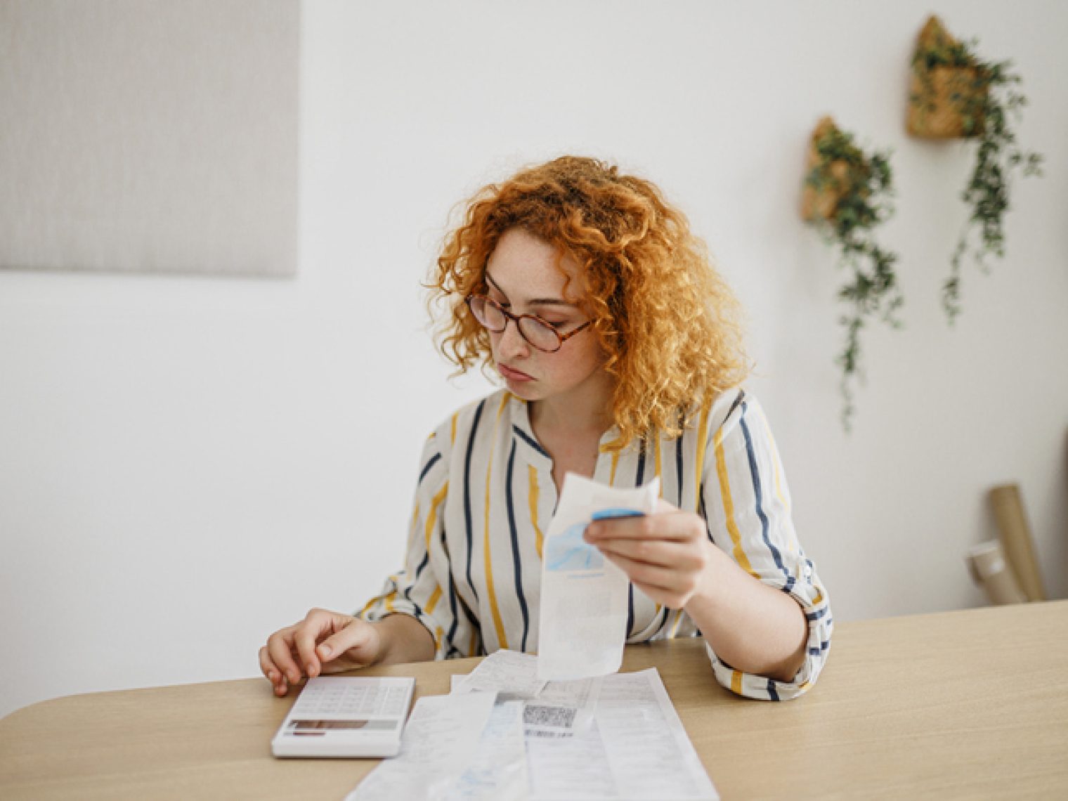 A focused young woman with curly hair and glasses examines receipts and documents spread out on a table.