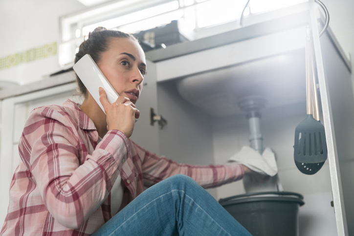 A concerned woman in a plaid shirt is talking on the phone while looking at a leaking sink with a bucket placed underneath.