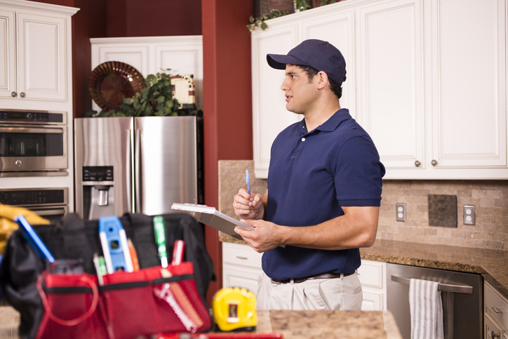 A home appraiser in a blue polo shirt and cap, holding a clipboard, attentively inspects a residential kitchen with a toolkit on the counter.