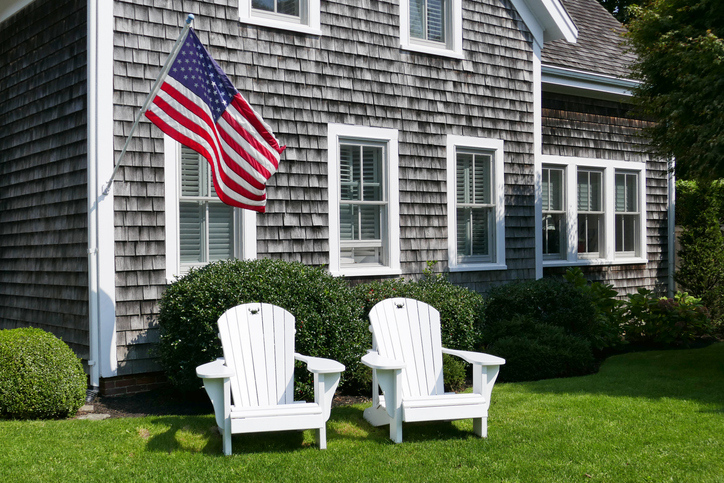 Two white Adirondack chairs on a well-kept lawn in front of a shingled home with an American flag displayed.