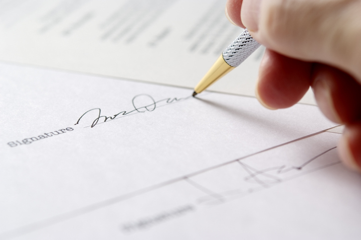 A close-up of a hand signing a formal document with a pen, indicating a signature being provided for a legal or official agreement.