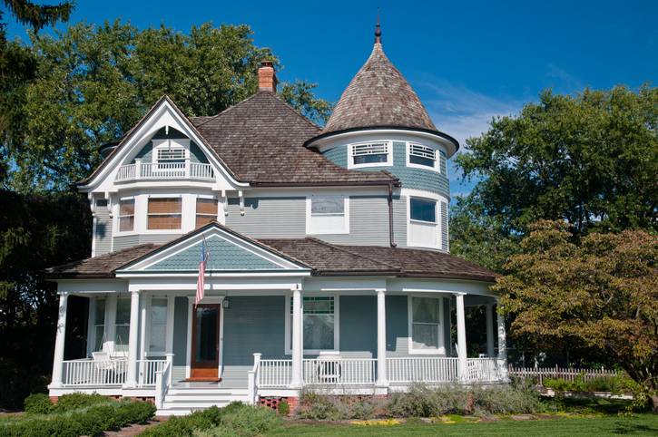 A Victorian-style house with a distinctive turret and wrap-around porch, set against a clear blue sky with surrounding greenery, exhibiting classic American architectural elements.