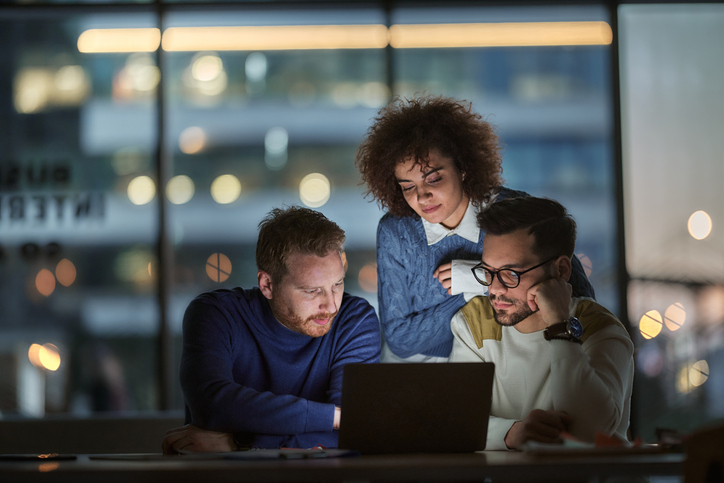 Three colleagues are focused on a laptop screen, working together in an office at night with city lights visible in the background through large windows.