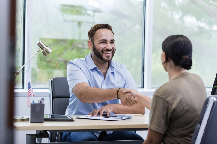 A smiling man extends a handshake across his desk to a woman in a bright office.